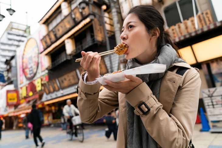 low angle shot of Asian Taiwanese female tourist biting into yummy takoyaki octopus ball on chopsticks at outdoor food street in Shinsekai Osaka Japan