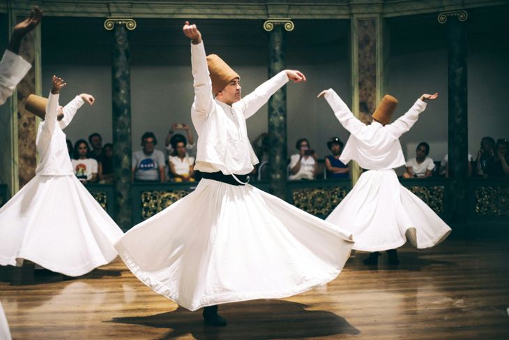 Three men wearing white traditional garments performing a whirling dance in a historical indoor setting.
