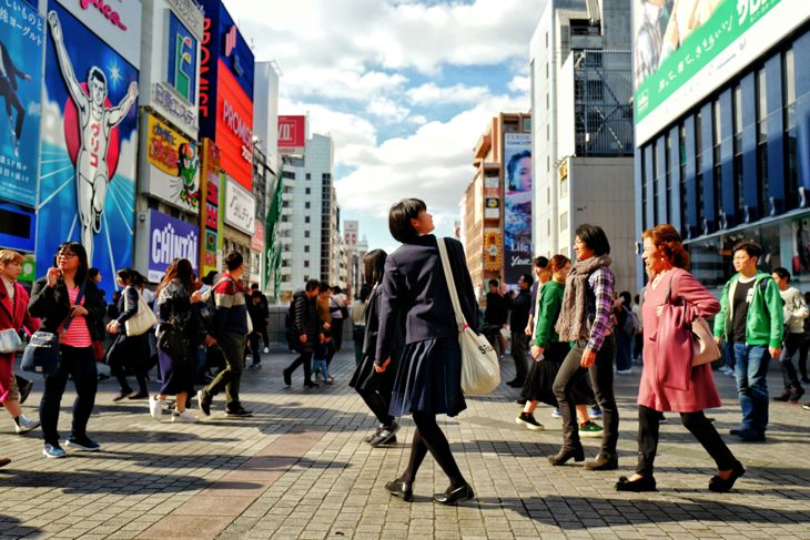 Busy urban street scene with people walking among tall buildings and colorful billboards under a blue sky.