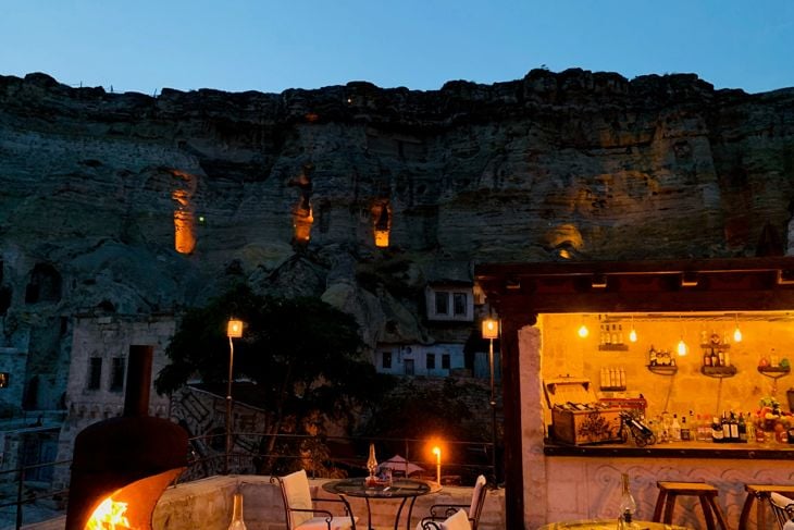 Outdoor seating area with tables and chairs lit by warm lights at dusk near rocky cliffs with cave openings.