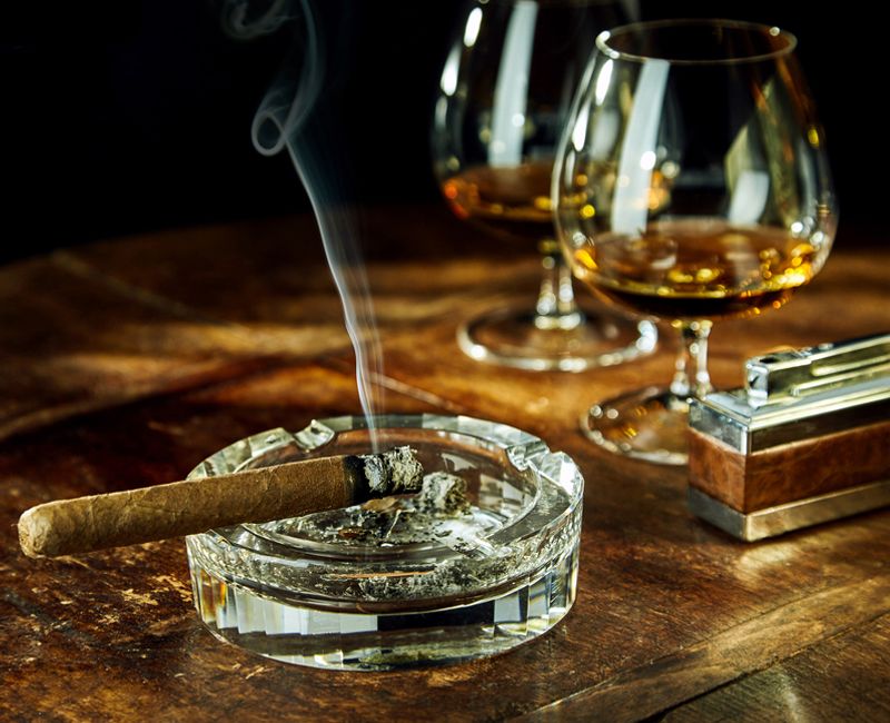 Glass ashtray with smoking cigar by two glasses filled with bourbon besides fancy lighter on a wood table