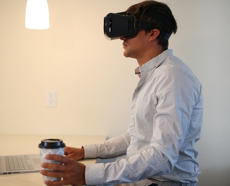 Businessman wearing virtual reality, augmented reality black glasses. Young man working on grey laptop, with cup of coffee. Inside the room, can be home or living room.