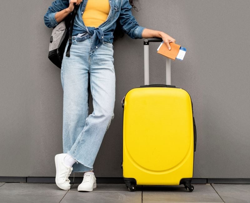 Unrecognizable woman in stylish casual outfit traveller standing over grey background, carrying yellow luggage and backpack, holding passport and flight tickets